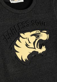 Sudadera gris con cuello acanalado, con un destacado gráfico de una cabeza de tigre amarilla y el texto "Alma Intrépida" en relieve arriba.