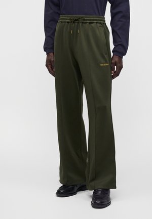 Mann trägt dunkelgrüne, weit geschnittene Hose mit Kordelzug in der Taille, goldener Schrift und schwarzen Schuhen, steht vor einem schlichten Hintergrund.