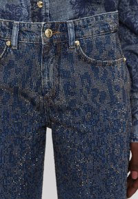 Denim jeans featuring a textured dark blue fabric with gold metallic accents and a patterned design. Front pockets visible with gold hardware.