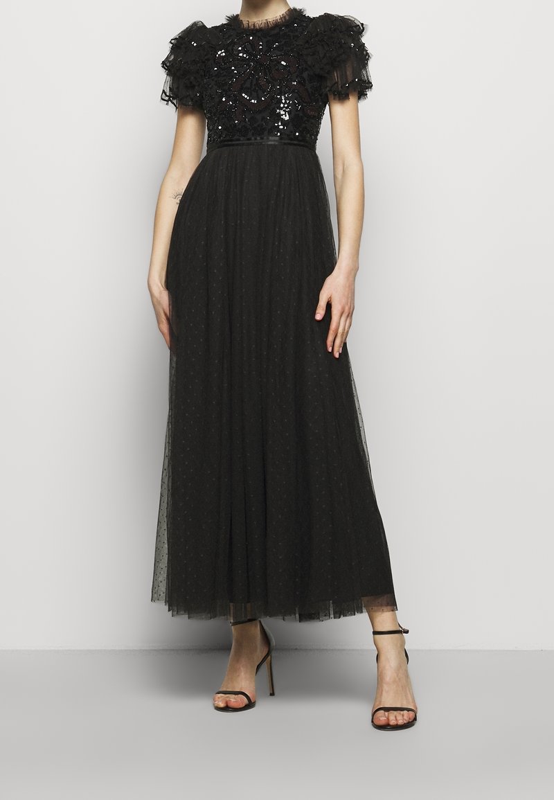 Robe noire longue jusqu'au sol avec corsage floral à sequins, manches courtes à volants, jupe en tulle transparent à pois, portée avec des talons noirs à bride cheville.