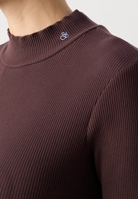 Brauner gerippter Rollkragenpullover mit hohem Kragen und einem hellblauen, gestickten Logo im Halsbereich. Weiche Textur und taillierte Passform.