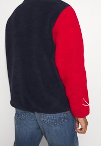 Fleecová bunda s tmavě modrým tělem a jasně červeným rukávem. Texturovaná tkanina s bílým akcentem na červeném rukávu. Ležérní střih.