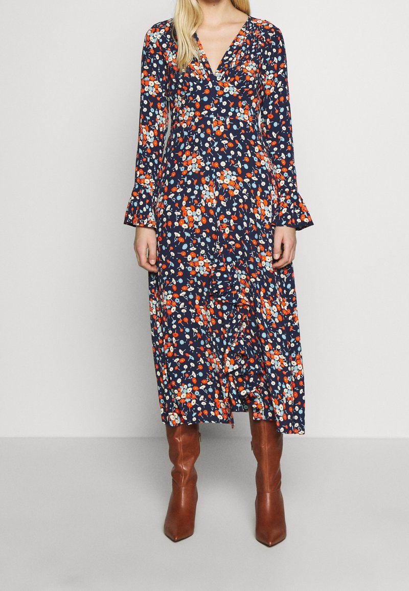 Robe portefeuille marine à fleurs avec manches longues, ornée de motifs floraux rouges et blancs et de volants aux manches, associée à des bottes marron à talons.