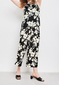 Černá květinová blůza s krémovými květy, výstřih ve tvaru klíčové dírky; černé široké kalhoty s květinovým vzorem. Modelka má na sobě černé sandály na podpatku.
