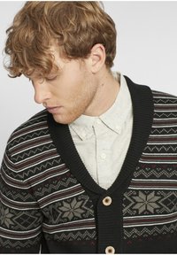 Svart cardigan med V-hals, med snøfnugg- og geometriske mønstre i grått, rødt og krem. Avsluttet med treknapper.