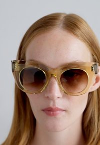 Lunettes de soleil surdimensionnées avec une monture transparente couleur ambre et des verres à dégradé brun. Accentuées par des détails géométriques sur les côtés.