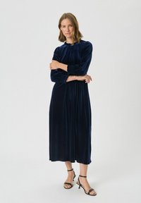 Marineblauwe fluwelen jurk met een hoge halslijn, pofmouwen en geplooide manchetten. Vloeiende midi-lengte met een gladde, rijke textuur.