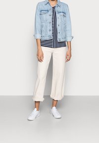 Helles blaues Jeansjacke über einem navyblauen und weißen gestreiften Hemd. Cremefarbene, hochgeschnittene Hosen mit umgeschlagenen Säumen, kombiniert mit weißen Sneakers.
