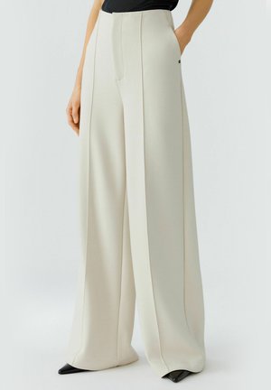 Pantalons taille haute, à jambes larges en tissu beige clair avec un devant plissé, poches latérales et chaussures noires pointues visibles en bas.