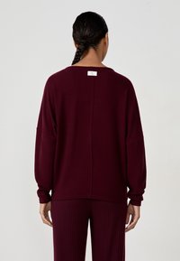 Burgundowy sweter o luźnym kroju, z opadającymi ramionami, prążkowanymi mankietami oraz centralnym szwem na plecach. Metka umieszczona przy dekolcie.