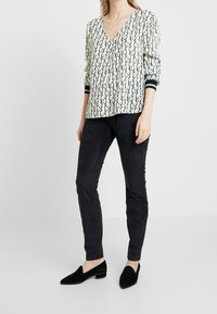 Biała bluzka z czarno-zielonym spiralnym wzorem, dekoltem w serek i obszernymi rękawami; zestawiona z czarnymi dopasowanymi spodniami i czarnymi loafersami.