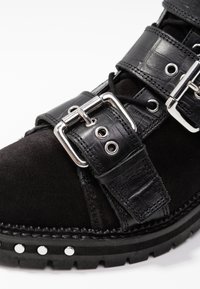 Botín negro de tobillo con cuero de ante y cuero con estampado de cocodrilo, equipado con hebillas y tachuelas plateadas, que destaca un diseño robusto.