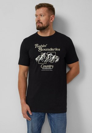 Baardige man die een zwart t-shirt draagt met bergafbeeldingen en de tekst "Pushin' Boundaries Country Pacemakers", staand tegen een grijze muur.