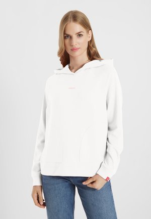 Jonge vrouw met een witte hoodie met zakken aan de voorkant en een blauwe spijkerbroek, staand tegen een effen lichtgrijze achtergrond.