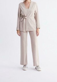 Beige stickad cardigan med knytmidja, matchad med vida byxor. Båda har en mjuk textur och avslappnad passform, kompletterat med vita sneakers.