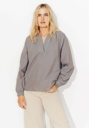 Femme blonde portant un chemisier à manches longues rayé bleu et marron et un pantalon beige, debout contre un fond blanc avec une main dans la poche.