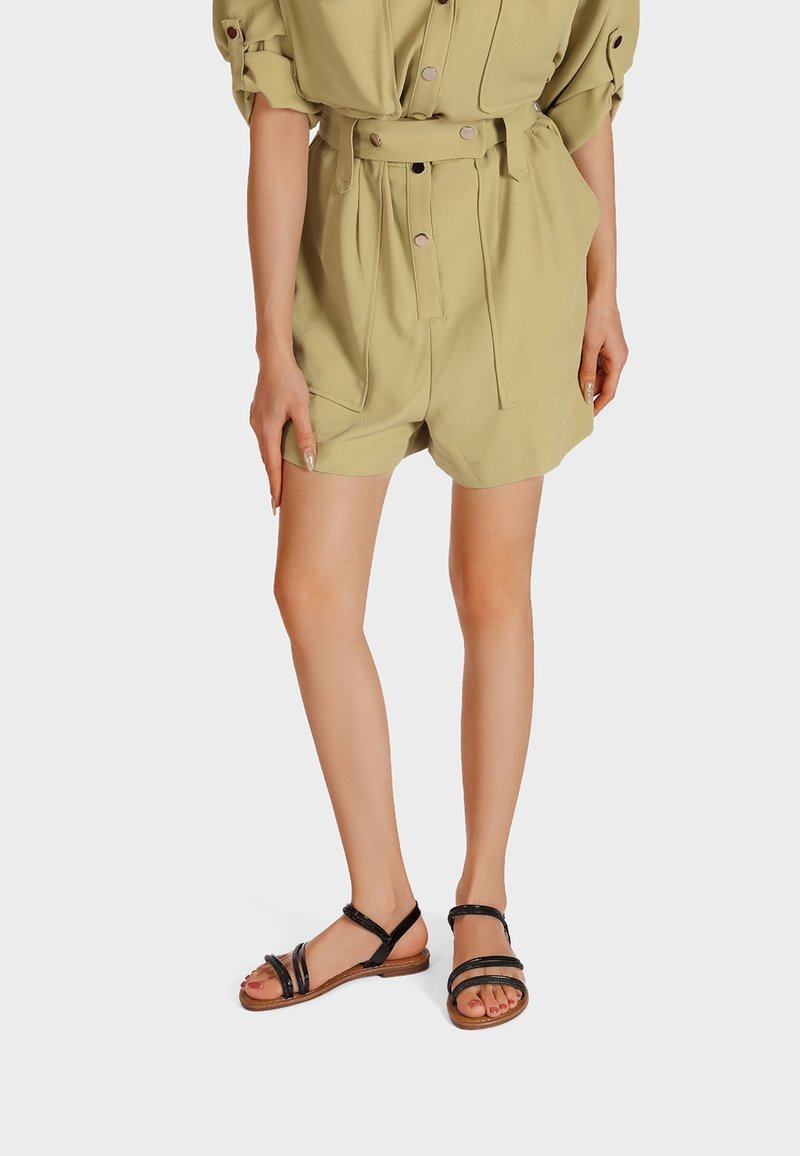 Sandales en cuir noir avec plusieurs fines lanières, semelle plate et accent marron à la base. Portées avec une tenue vert olive clair.