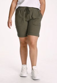 Pantaloni corti verde oliva con orlo arrotolato e grandi tasche, realizzati in tessuto leggero, abbinati a sneakers bianche da ginnastica.