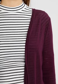 Burgundy kofta med öppen front, lager på lager över en svart-vitt randig polotröja. Tyget ser mjukt ut med en lätt struktur.