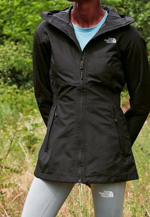 Persona con una chaqueta negra de The North Face sobre una camisa azul clara y leggings grises claros de The North Face al aire libre, en un entorno natural verde.
