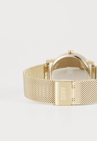 Orologio con cinturino in rete dorata e cassa rotonda, dotato di una finitura metallica e di una chiusura incisa con "DKNY". Design minimalista con quadrante chiaro.