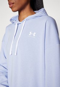 Svijetloplava dukserica s mekanom teksturom, podesivim bijelim žnjorcima i kontrastnim bijelim Under Armour logom na prsima.