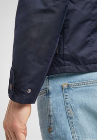 Marinblå nylonjacka med en tryckknapp i mudd och texturerad yta, kombinerad med ljusblå denimjeans med synliga sömmar.