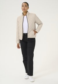 Veste matelassée beige avec un col haut, une fermeture éclair noire et des poches latérales, associée à un pantalon noir et des baskets blanches. Design simple et fonctionnel.