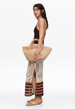 Mujer con un top sin mangas marrón oscuro, falda larga de crochet con abertura, sosteniendo un gran bolso de paja tejido, lleva sandalias amarillas, está de perfil.