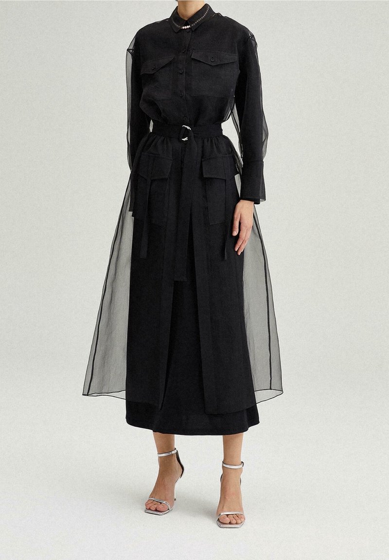 Femme portant une longue robe noire ceinturée avec des manches longues transparentes et des poches, associée à des sandales argentées à talons hauts avec bride à la cheville.