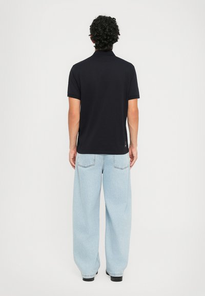 Man med lockigt hår, iklädd en svart kortärmad polo och ljusblå vida jeans, står vänd bortåt mot en vit bakgrund.