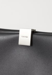 Sac à main en cuir noir avec une surface texturée et un fermoir carré en métal argenté gravé "Calvin Klein."