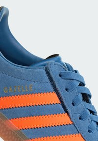 Zilas zamšs sporta apavi ar trim oranžām svītrām, zelta "Gazelle" logotipu, apaļām šņorēm un teksturētu gumijas zoli. Tuvplāna skats.
