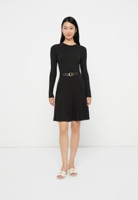 Donna che indossa un abito di maglia nero a maniche lunghe con una cintura nera e tacchi bianchi aperti, in piedi contro uno sfondo bianco semplice.