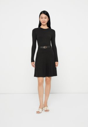 Donna che indossa un abito di maglia nero a maniche lunghe con una cintura nera e tacchi bianchi aperti, in piedi contro uno sfondo bianco semplice.