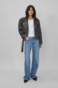 Moteris stovi prieš pilką foną, dėvi juodą odinę striukę, baltą palaidinę, mėlynas platėjantį juosmenį džinsines kelnes ir juodus smailėjančius batus.