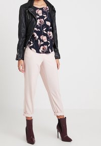 Giacca di pelle nera sopra una blusa floreale blu navy, abbinata a pantaloni tapered rosa chiaro e stivaletti alla caviglia bordeaux scuro con design a fessura.