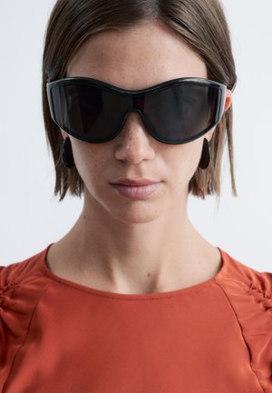 Mujer con cabello corto castaño que lleva gafas de sol negras de gran tamaño, pendientes de aro negros y una blusa naranja sobre un fondo blanco.