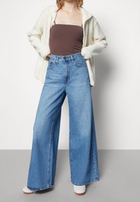 Vidvuxna blå jeans med hög midja, kombinerade med ett brunt linne och en fluffig cremefärgad zip-up-jacka. Looken kompletteras med vita skor.