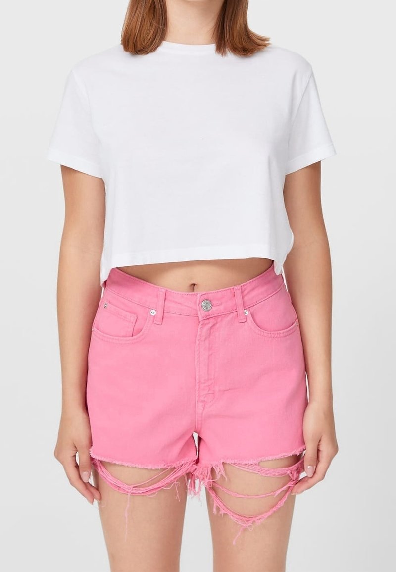 Personne portant un t-shirt court blanc et un short en jean rose taille haute avec des ourlets abîmés et des bretelles effilochées.