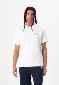 Weißes Baumwoll-T-Shirt mit einem mintgrünen Nike SB-Logo auf der Brust. Mit kurzen Ärmeln und einem lockeren Schnitt, getragen mit navyblauen Hosen.