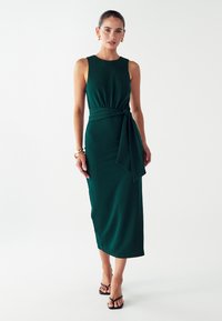 Duga, bezrukava zelena haljina od glatkog materijala, s vezanim akcentom u struku i midi dužinom. Manekenka nosi crne sandale.