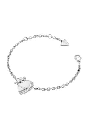 Bracelet chaîne en argent avec un charm en forme de cœur gravé "GUESS" et de petits cristaux le long d'un bord, fermoir et étiquette de marque triangulaire attachée.