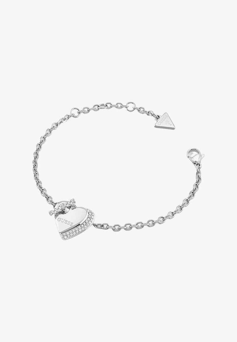Bracelet chaîne en argent avec un charm en forme de cœur gravé "GUESS" et de petits cristaux le long d'un bord, fermoir et étiquette de marque triangulaire attachée.