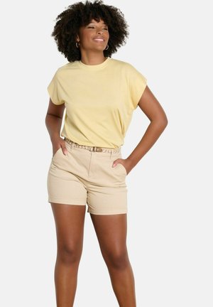 Mujer con cabello rizado que lleva una blusa sin mangas de color amarillo claro y pantalones cortos beige con un cinturón trenzado, con las manos en los bolsillos, sonriendo.
