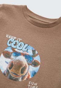 Close-up van een bruine shirt met een graphic van een eland met een zonnebril en sneeuw, met de tekst "Hou het koel" en "Leuke tijden."