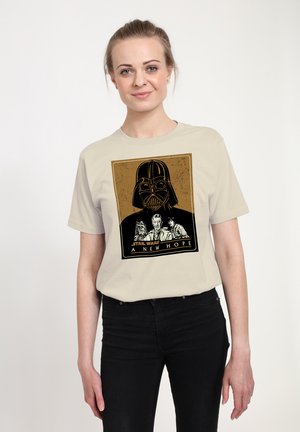 Usmívající se žena v béžovém tričku Star Wars s helmou Darth Vadera a postavami Chewbacca, Obi-Wan a Luke Skywalker s nápisem "Nová naděje".