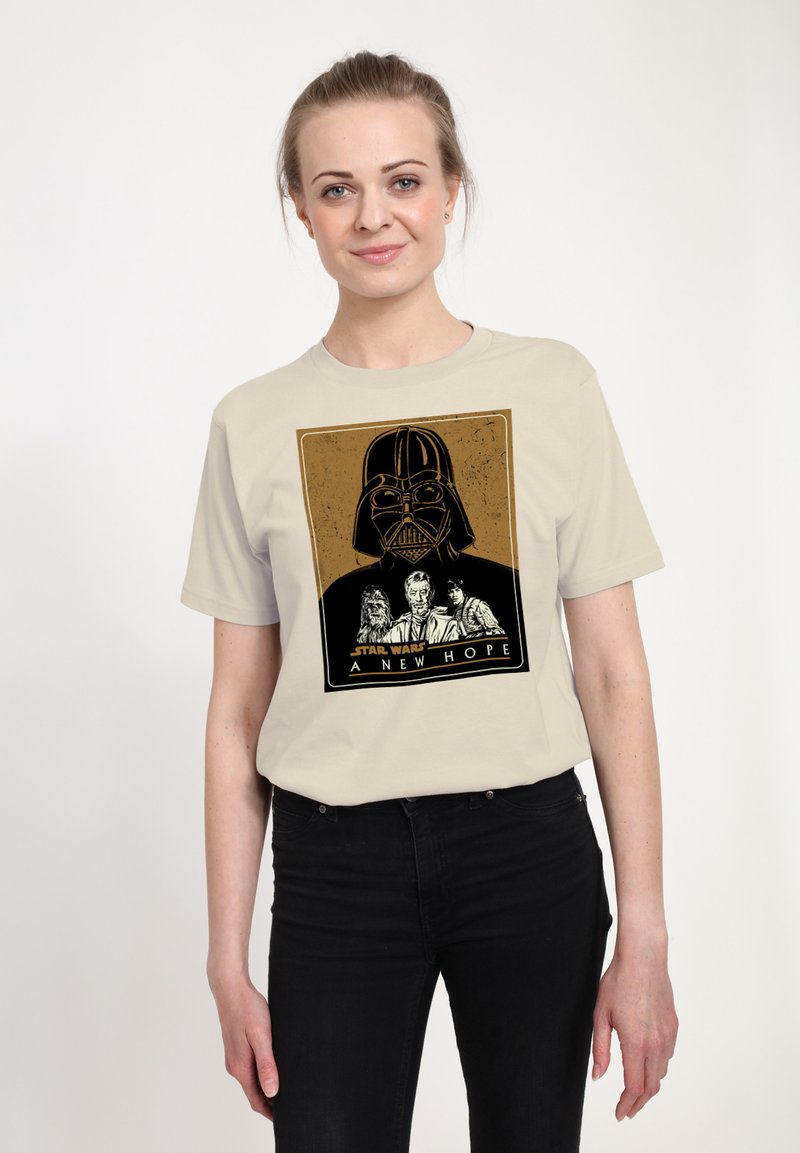 Usmívající se žena v béžovém tričku Star Wars s helmou Darth Vadera a postavami Chewbacca, Obi-Wan a Luke Skywalker s nápisem "Nová naděje".