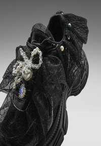Μαύρο αθλητικό παπούτσι διακοσμημένο με μαύρο δαντελένιο ύφασμα, διακοσμητικά από μαργαριτάρια και κρυσταλλικούς χάντρες και ένα μπλε λίθο στη δαντέλα κοντά στο μπροστινό μέρος.