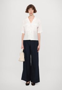 Witte, kortlopende linnen blouse met strikdetail, gecombineerd met wijde marineblauwe broek en een kleine beige handtas. Toont een eenvoudig, modern ontwerp.